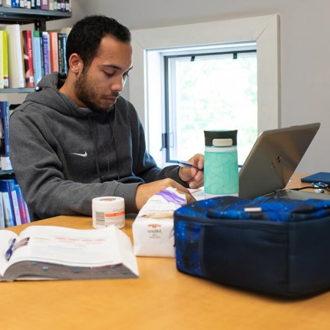 A male student studies in the library with textbooks and a laptop